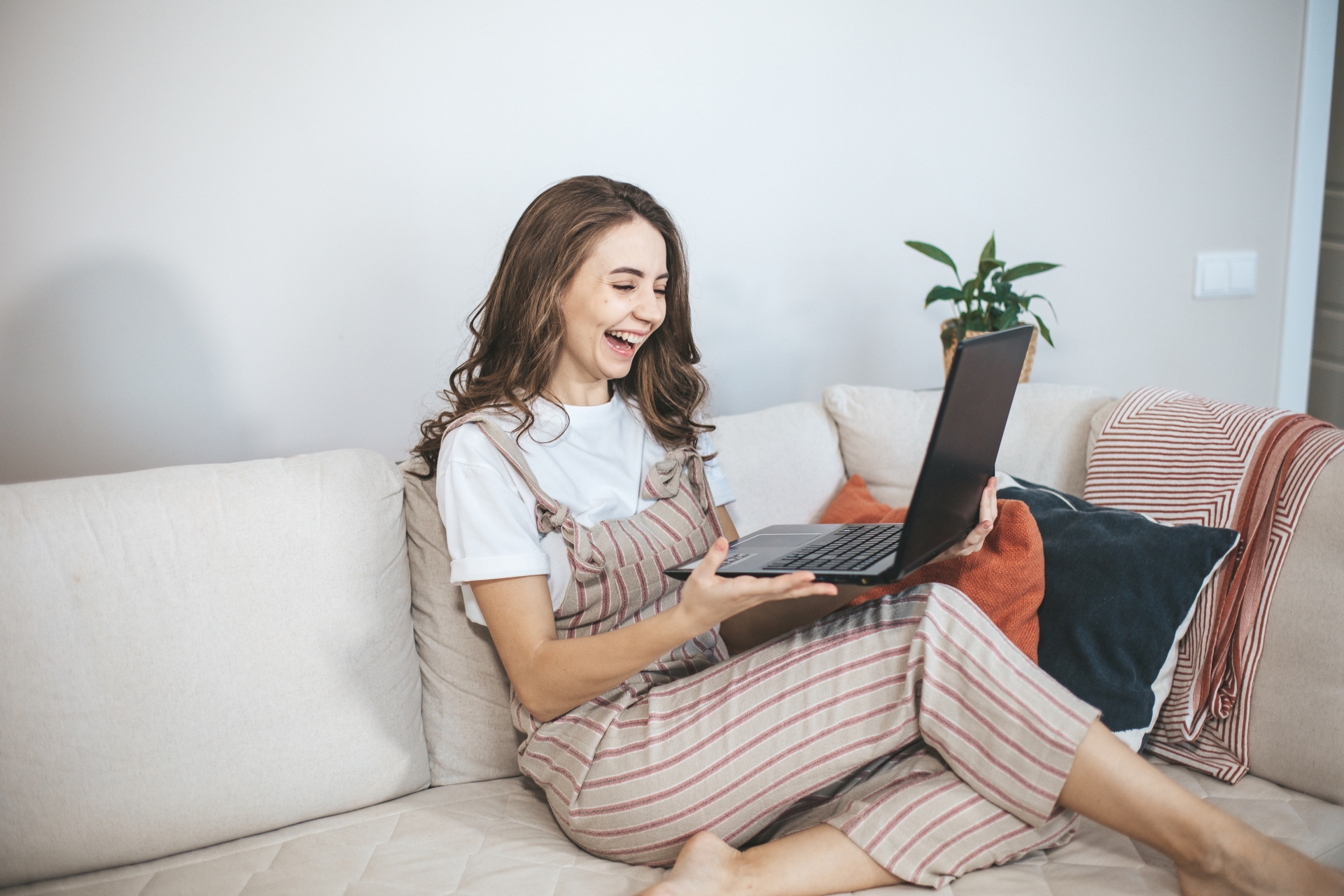 Joyful young woman laughing and looking at a laptop on a cozy couch.