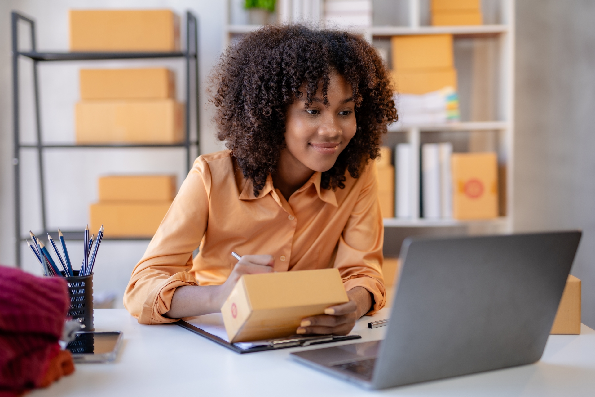 Smiling woman with curly hair using laptop and holding a parcel in an office with shelves of packages.